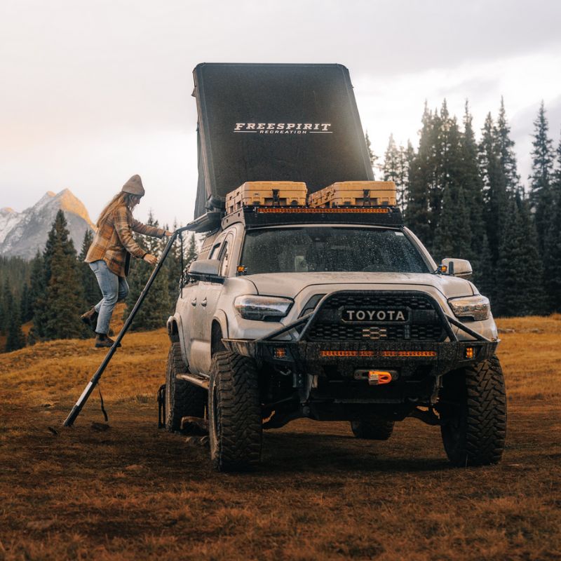 freespirit-recreation-aspen-v2-standard-hard-shell-roof-top-tent-black-open-front-view-on-toyota-tacoma-with-woman-climbing-ladder-in-nature