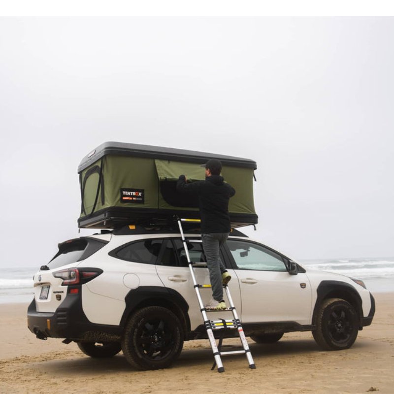 tentbox-classic-2-hard-shell-roof-top-tent-forest-green-open-front-view-on-subaru-outback-with-person-on-ladder-at-the-beach